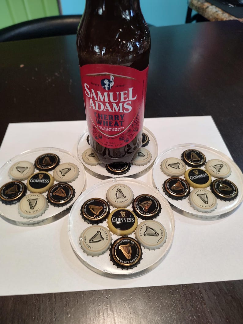May include: A bottle of Samuel Adams Cherry Wheat beer rests on a clear coaster surrounded by other coasters. Each coaster is decorated with beer bottle caps, including Guinness caps. The coasters are on a dark surface.