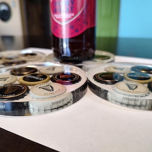 May include: Circular coasters with embedded beer bottle caps. The caps feature various logos and designs, including the Guinness harp. The coasters are clear with a black and white border, and a bottle rests on one.