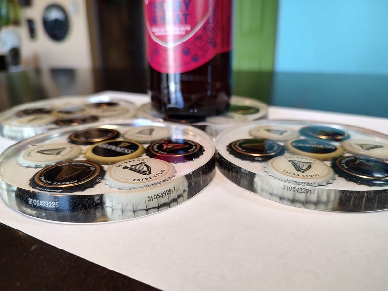 May include: Circular coasters with embedded beer bottle caps. The caps feature various logos and designs, including the Guinness harp. The coasters are clear with a black and white border, and a bottle rests on one.