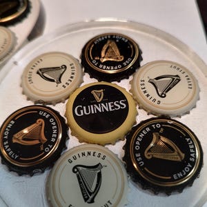 May include: A collection of Guinness bottle caps, featuring the iconic harp logo and text like "Extra Stout" and "Enjoy Safely." The caps are black, gold, and cream-colored, arranged on a clear surface.