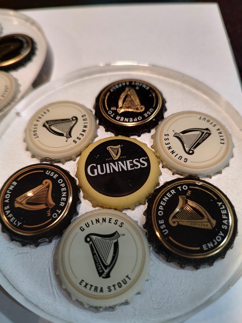 May include: A collection of Guinness bottle caps, featuring the iconic harp logo and text like "Extra Stout" and "Enjoy Safely." The caps are black, gold, and cream-colored, arranged on a clear surface.