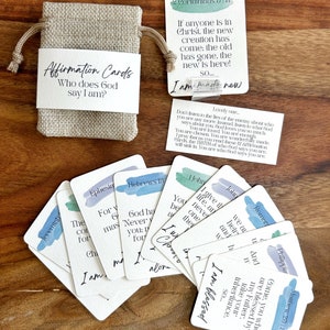 May include: A set of 12 affirmation cards featuring Bible verses printed on them. The cards are decorated with watercolour-like backgrounds in shades of blue and green. The cards are arranged in a fan shape on a wooden surface. The text on the cards includes verses from the Bible, such as "If anyone is in Christ, the new creation has come: the old has gone, the new is here! So... I am made new." and "Come, you who are blessed by my Father, inherit your inheritance. So... I am blessed."