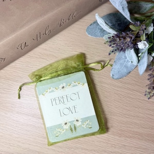 May include: A rectangular card with the text PERFECT LOVE printed in black, decorated with white floral borders on a light green background, enclosed in a sheer olive green organza drawstring pouch with a satin ribbon closure.