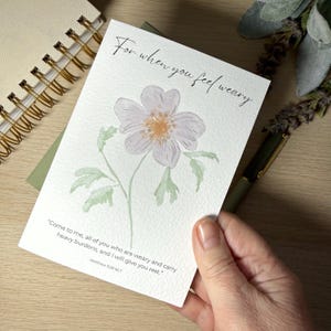 May include: A watercolour greeting card with a light purple flower and a yellow centre, accompanied by green leaves. The card displays the text "For when you feel weary" and a Bible verse. The card is held in a hand.