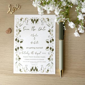May include: A wedding invitation with the text "Save the Date" and the names "Charlie & Arabella". The invitation is decorated with a floral design and a pen rests beside it. The date is 8th August 2026.