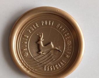 May include: A round gold wax seal featuring a central embossed reindeer leaping over horizontal lines. The text reads NORTH POLE POST OFFICE OFFICIAL MAIL, with small snowflake icons separating the words. The seal has a smooth, raised border.