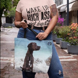 Pode incluir: Uma camiseta rosa com o texto "Wake Up Kick Ass Pet Dogs" em letras pretas. A pessoa está segurando uma bolsa marrom com um cachorro marrom nela. O cachorro está sentado em uma praia com pedras e água ao fundo.