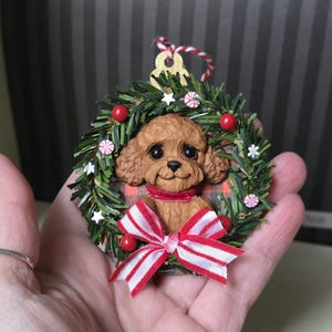 May include: A Christmas ornament featuring a brown poodle dog inside a green wreath decorated with red berries, white stars, and peppermint candies. The dog wears a red scarf and a red and white striped bow. A gold-colored tag and red and white string are at the top.