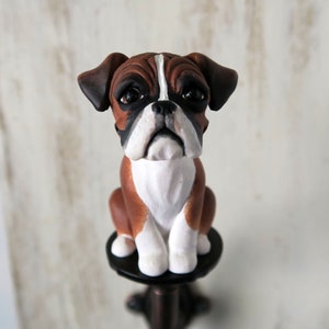 May include: A brown and white boxer dog figurine with a black nose and brown eyes. The figurine is sitting on a black pedestal.