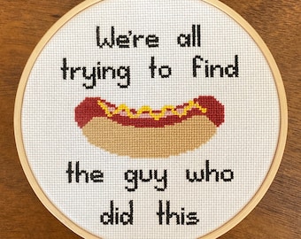 May include: Embroidered hoop art with the text "We're all trying to find the guy who did this" and a cartoon hot dog with mustard and ketchup.