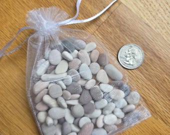 May include: A small, sheer white mesh bag filled with smooth, assorted-color river rocks. The rocks are primarily gray, white, and pink, with a few green ones. A US quarter coin is next to the bag for size comparison.