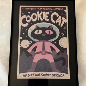 May include: Framed print featuring a cartoon cat in a spacesuit, with the text "Cookie Cat" and "A Refugee of an Interstellar War." The cat is black with pink accents, set against a space-themed background with planets and stars.