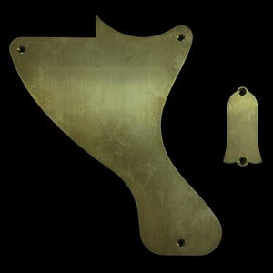 May include: Two brass guitar parts on a black background. The larger piece has a unique shape with three screw holes. The smaller piece is bell-shaped with two screw holes. Both pieces have a brushed metal finish.