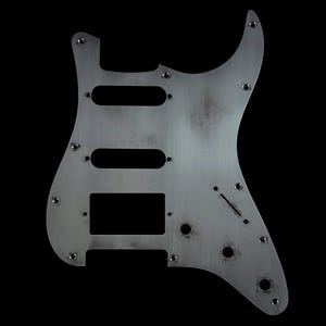 Könnte beinhalten: Ein silberfarbenes Gitarren-Schlagbrett mit drei rechteckigen Aussparungen für Tonabnehmer und mehreren Schraubenlöchern. Das Schlagbrett hat eine verwitterte, gebürstete Metalloberfläche. Es ist für eine E-Gitarre konzipiert und vor einem schwarzen Hintergrund platziert.