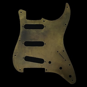 May include: A brass-finished guitar pickguard with an aged appearance. It features three elongated slots for pickups, multiple screw holes, and a distinctive shape. The pickguard is set against a black background, showing signs of wear.