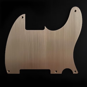 Peut inclure: Un pickguard de guitare brossé de couleur laiton avec un motif de grain vertical. Le pickguard a une forme distinctive avec des bords arrondis et angulaires, et plusieurs trous pré-percés pour le montage. Le fond est noir uni.