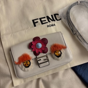 Può includere: Una borsa in pelle bianca con chiusura in metallo argentato Fendi Roma, una grande decorazione floreale bordeaux e rosa, e due dettagli a forma di occhio con bordo in pelliccia arancione e centri in cristallo color ambra, con sacca protettiva.