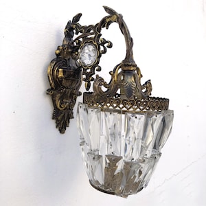 May include: An antique wall sconce with a bronze finish and a clear crystal shade. The sconce features ornate detailing, including a decorative backplate, a crystal accent, and a curved arm. The shade is made of faceted crystals.