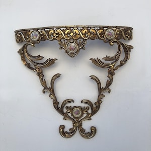 Puede incluir: Un estante de pared con un marco de metal dorado ornamentado con intrincados detalles de volutas y filigrana. El estante está acentuado con cinco pequeñas incrustaciones circulares de porcelana, cada una decorada con un colorido patrón floral.