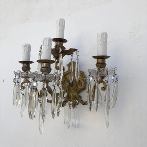 May include: A wall-mounted light fixture featuring a gold-toned metal frame with ornate scrollwork. It holds four white candle-shaped light covers, each surrounded by clear, faceted glass crystal prisms that hang from scalloped glass bobeches.