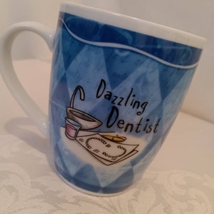 May include: A white ceramic mug with a blue and white diamond pattern. The mug has a handle and says "Dazzling Dentist" in black lettering. The mug has a drawing of a sink, a glass of water, a toothbrush, and a tray of teeth.