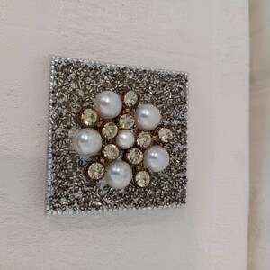 May include: A square-shaped wall decor with a silver background and a cluster of white pearls and rhinestones in the center.
