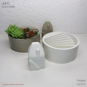 Könnte beinhalten: Drei Beton-Pflanzkübel in verschiedenen Formen und Größen. Der größte Kübel ist weiß und rund mit einem gewellten Design. Der mittlere Kübel ist grau und rund. Der kleinste Kübel ist weiß und hat die Form eines Hauses. Die Kübel stehen auf einer weißen Oberfläche. Art & Concrete Sayfiye Portland.