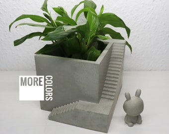 May include: A concrete planter with a built-in staircase design. The planter is filled with a green plant and has the words "MORE COLORS" printed on the side. A small gray rabbit figurine is in the foreground.
