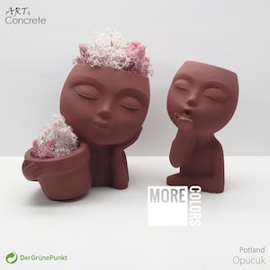 Puede incluir: Dos macetas de cerámica de color terracota con forma de rostro humano. Una maceta tiene los ojos cerrados y sostiene una maceta más pequeña con musgo rosa y blanco. La otra maceta tiene los ojos cerrados y está dando un beso. El texto "MORE COLORS" está impreso en blanco sobre un cuadrado negro.