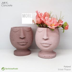 Könnte beinhalten: Zwei rosafarbene Beton-Gesichts-Pflanzer mit geschlossenen Augen und einem lächelnden Mund. Der Pflanzer rechts hat rosa Blumen drin.