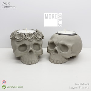Könnte beinhalten: Zwei graue Beton-Schädel-Kerzenhalter. Ein Schädel hat einen Blumenkranz um die Oberseite. Der Text "MORE COLORS" befindet sich in einem weißen Quadrat in der oberen rechten Ecke des Bildes.