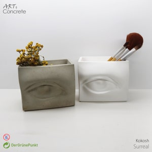 May include: Two concrete planters shaped like a human eye, one in gray and one in white. The gray planter has dried yellow flowers inside. The white planter has a set of makeup brushes inside.
