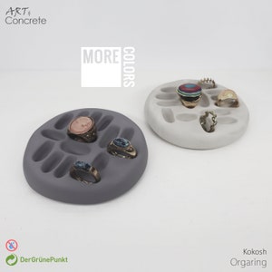 May include: Two concrete ring dishes, one grey and one white, with multiple ring-shaped indentations. The grey dish has three rings in it, and the white dish has five rings and a bracelet. The text "MORE COLORS" is printed in black on a white background above the dishes.