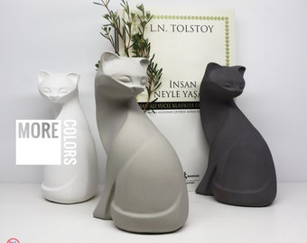 May include: Three concrete cat figurines in white, gray, and black. The figurines are sitting on a white surface. The text "More Colors" is visible in the image. The text "DecoDomDom Le Chat Noir" is also visible in the image.