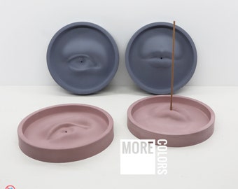 Puede incluir: Soportes de incienso de hormigón en gris y rosa, cada uno con un diseño de ojo o boca esculpido. Un soporte tiene una varilla de incienso. El texto "ART & Concrete" y "MORE COLORS" son visibles.