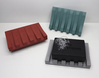 May include: Three concrete soap dishes in red, teal, and gray. The red and teal dishes have a ridged design. A gray dish holds a black bar of soap with bubbles. The text "Art & Concrete" and "Pirupak Sobi Flobi" are visible.
