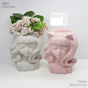 Può includere: Due vasi da fiori in cemento a forma di testa di ragazza con capelli lunghi e orecchie da gatto. Il vaso a sinistra è grigio e il vaso a destra è rosa. Il testo "MORE COLORS" è visibile sullo sfondo.