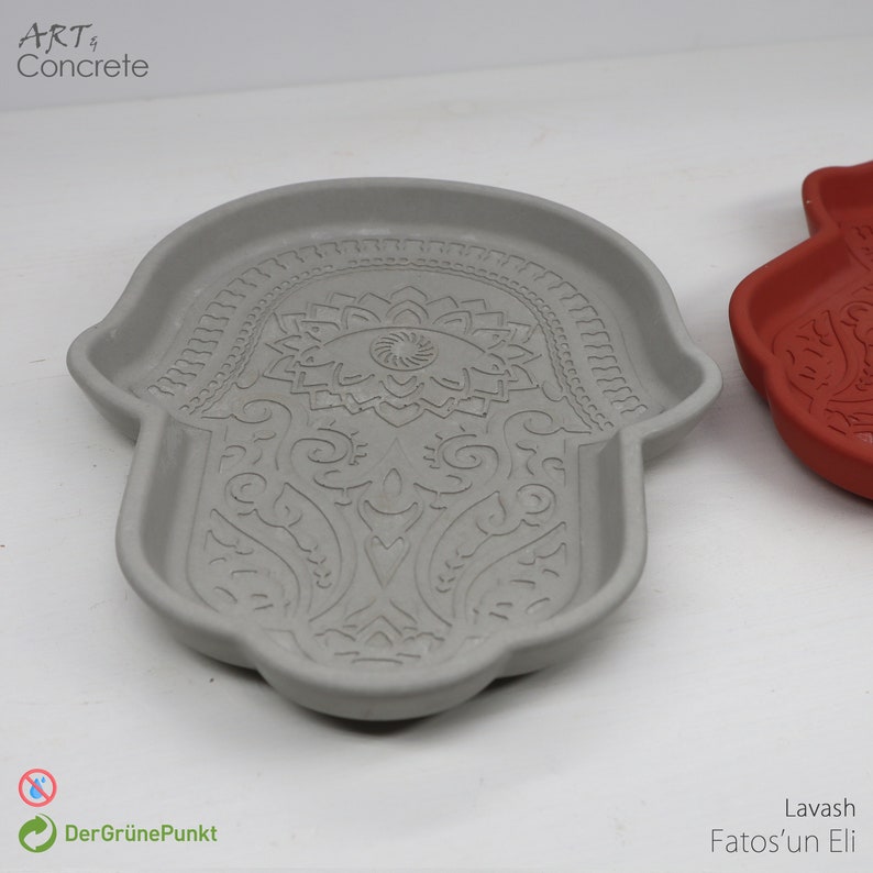 May include: A gray concrete tray shaped like a hamsa hand with an intricate floral design. The tray is a decorative item for home decor.