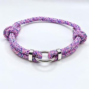 May include: A circular braided rope collar featuring a multi-colored pattern of pink, purple, and light blue threads. The design includes two silver-toned metal sliding clasps and a central silver-toned metal ring, with two decorative knots.