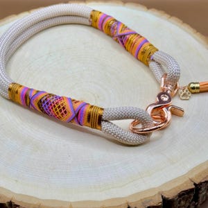May include: A beige rope dog collar with a colorful woven design and a rose gold clasp. The collar has a small rose gold charm with a paw print.
