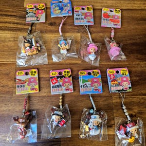 Puede incluir: Una colección de 10 amuletos de personajes de anime coloridos con diferentes diseños. Cada amuleto está unido a una cuerda y tiene una pequeña tarjeta con texto japonés. Los amuletos representan personajes de la popular serie de anime One Piece.