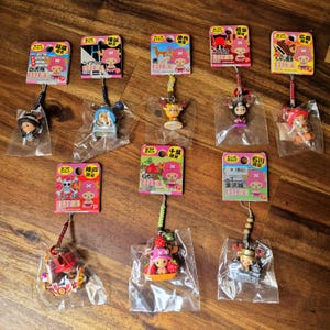 May include: A collection of 10 colourful One Piece character charms. Each charm is packaged in a clear plastic bag with a small card featuring the character's name and location. The charms are made of plastic and feature various characters from the anime series, including Luffy, Zoro, and Sanji.