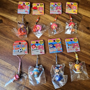 Puede incluir: Una colección de 10 amuletos de personajes de anime coloridos con diferentes diseños. Cada amuleto está unido a una cuerda y empaquetado en una bolsa de plástico transparente. Los amuletos representan personajes de la popular serie de anime One Piece.
