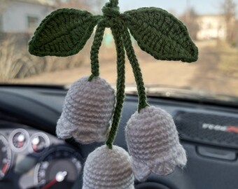May include: A white crocheted flower garland with three white bells and two green leaves hanging from a rearview mirror in a car.
