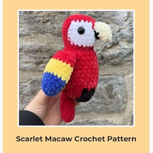 May include: A crocheted scarlet macaw parrot toy with a yellow, blue, and red wing. The parrot has a white face with a black eye and a yellow beak.