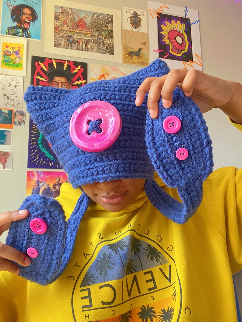 May include: A blue crocheted hat with a pink button and a matching blue crocheted scarf with pink buttons. The hat and scarf are worn by a person wearing a yellow sweatshirt with a graphic of palm trees and the text "VENICE" on the front.