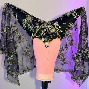 May include: A black headdress featuring a pointed, V-shaped structure adorned with intricate gold and silver floral embroidery. The piece includes sheer black lace side panels with matching metallic stitching, a central white pearl, and three small blue beads.