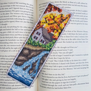 May include: A cross-stitch bookmark depicting a small house with a blue roof and a waterfall in front of it. The house is surrounded by green trees and a yellow, orange, and red autumnal tree. The bookmark is made of white fabric with a scalloped edge.