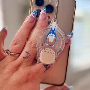 May include: A clear circular phone grip featuring a stacked graphic of three Totoro characters. The bottom character is large and beige, the middle is blue, and the top is small and white. The grip is attached to the back of a gold-toned smartphone.