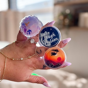 May include: A collection of round, decorative resin art pieces. One features a sunset scene with a silhouette, another has a celestial design, and a third displays the text "High Loon" with a floral illustration. The pieces are held in a hand.